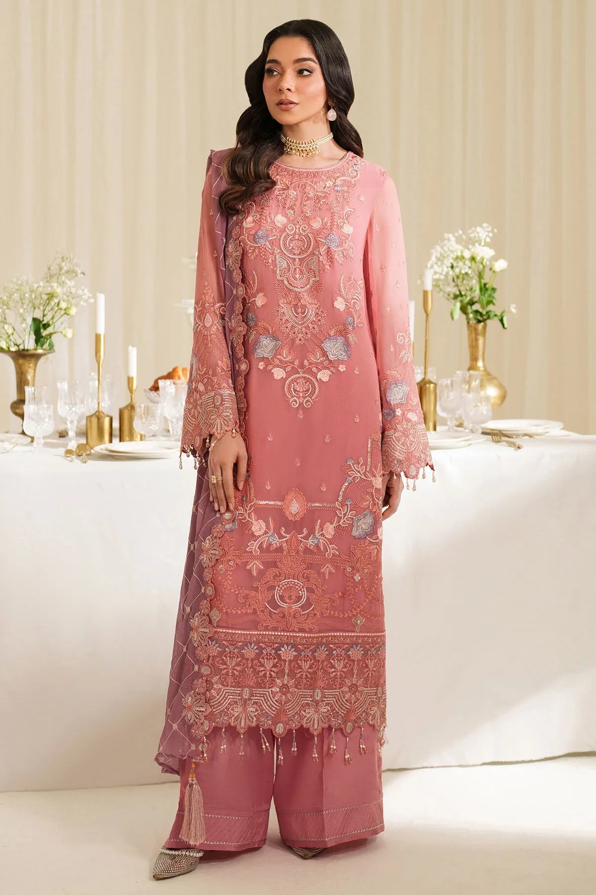 Woman in a pink embroidered outfit standing in a decorated room with flowers and candles.