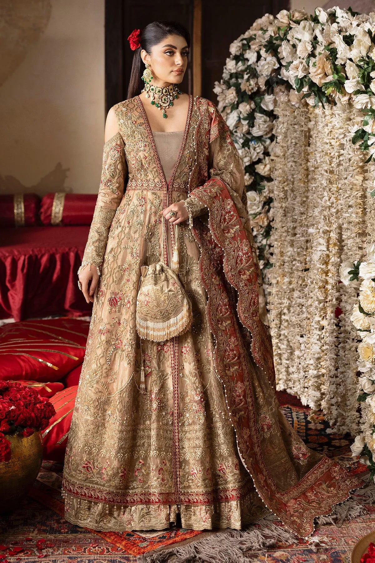 A woman wearing a traditional, embroidered beige gown with a floral pattern, paired with a dupatta and organza sleeves, standing in a decorative setting with floral arrangements in the background.