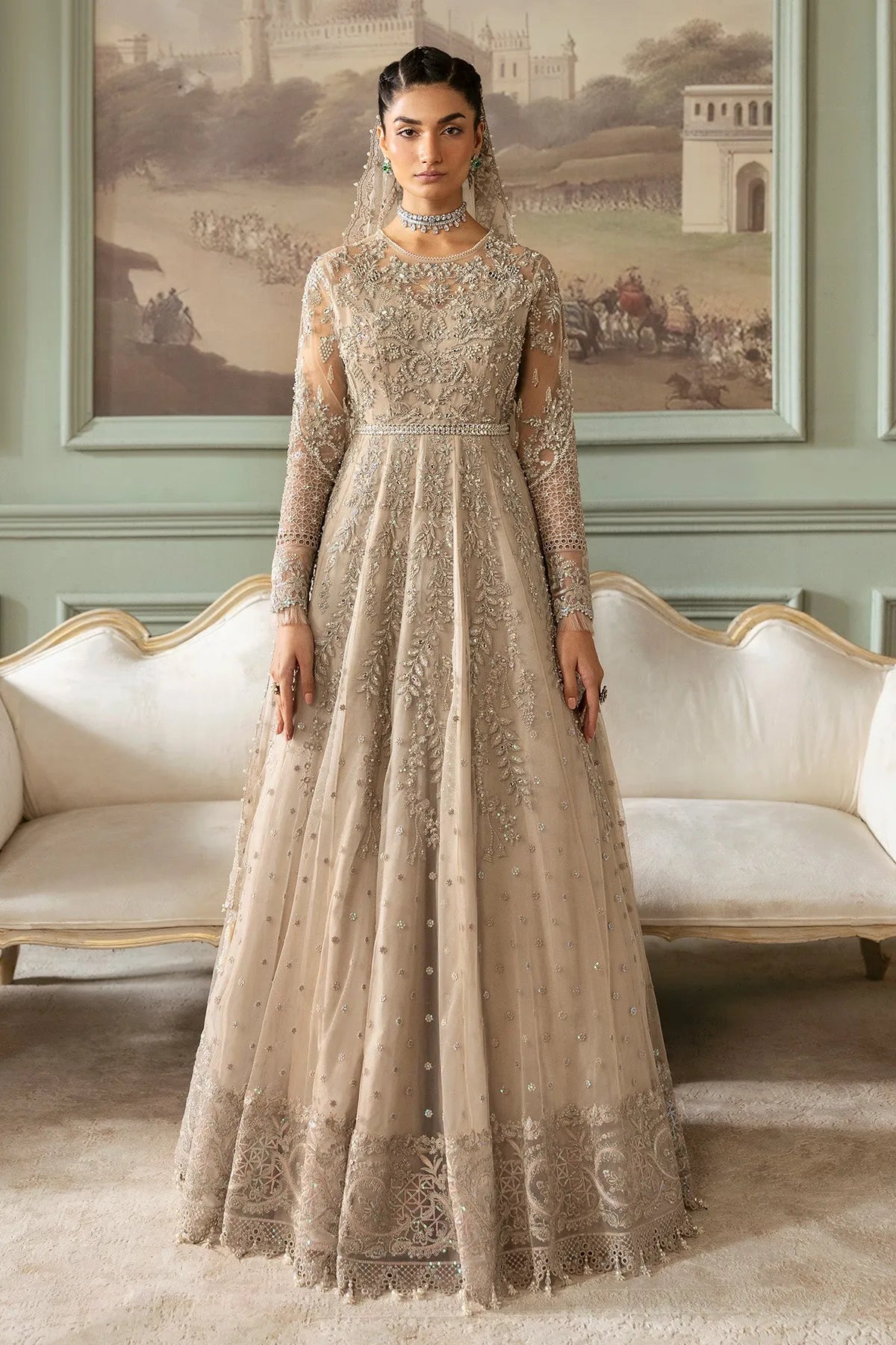 A woman wearing a beige and silver embroidered gown with long sleeves and a floor-length skirt, accessorized with a net dupatta.