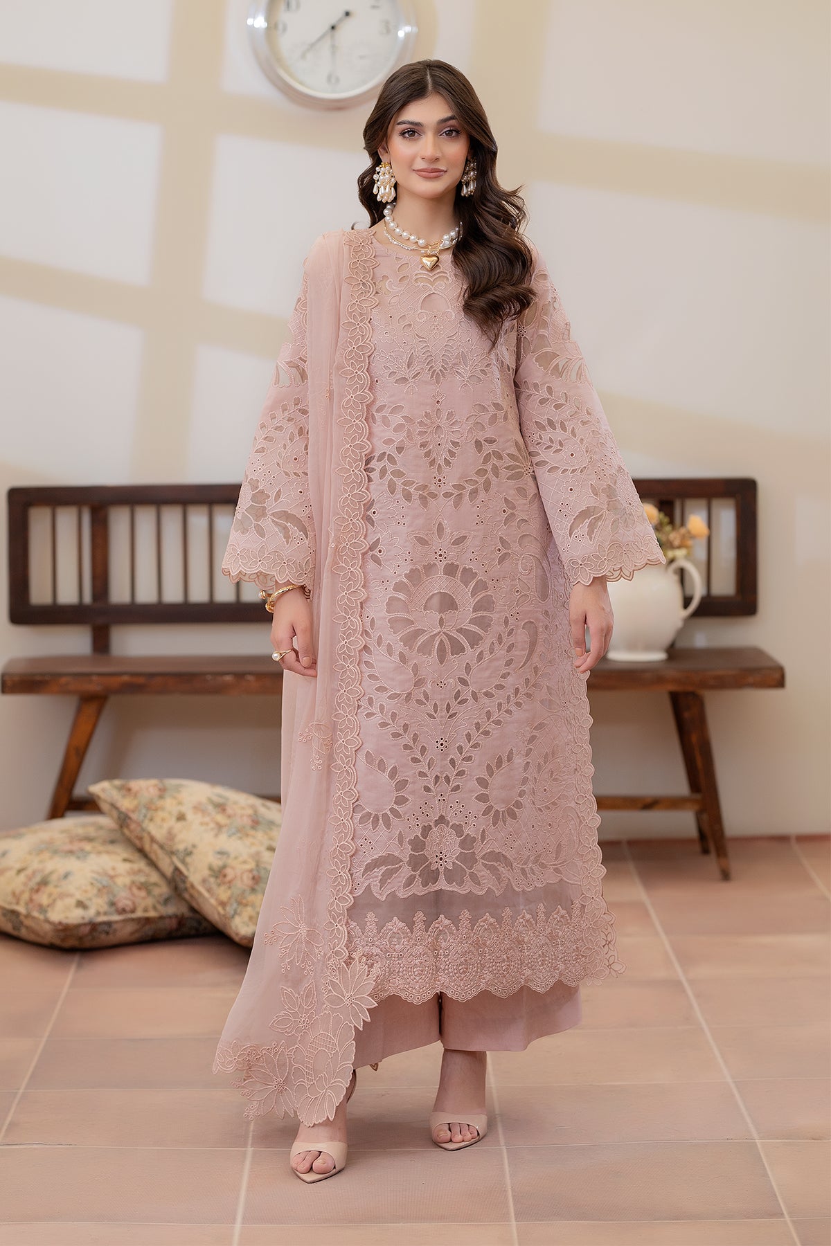 Woman wearing a pink embroidered outfit in a room with wooden furniture and a clock on the wall.