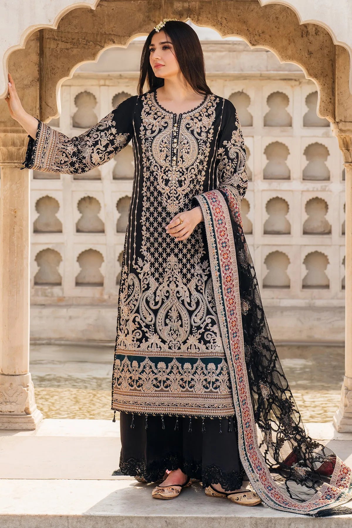 A woman wearing a black, red, and green embroidered three-piece clothing ensemble, consisting of a net shirt with plain back and sleeves, a net dupatta, and raw silk trousers, standing against an archway background.