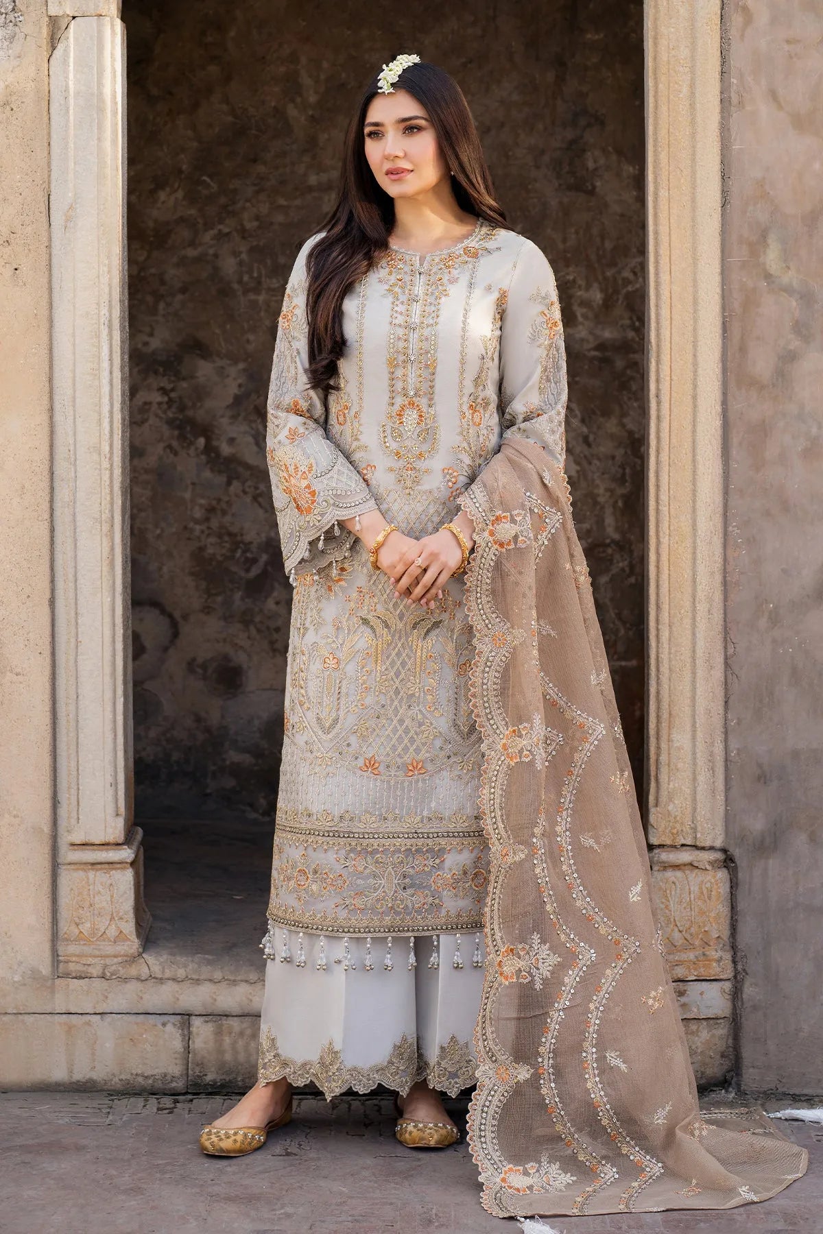 A woman wearing a traditional three-piece Mushk outfit with embroidered organza front and plain organza back with a dupatta. The outfit is in shades of gray and beige with intricate embroidery.