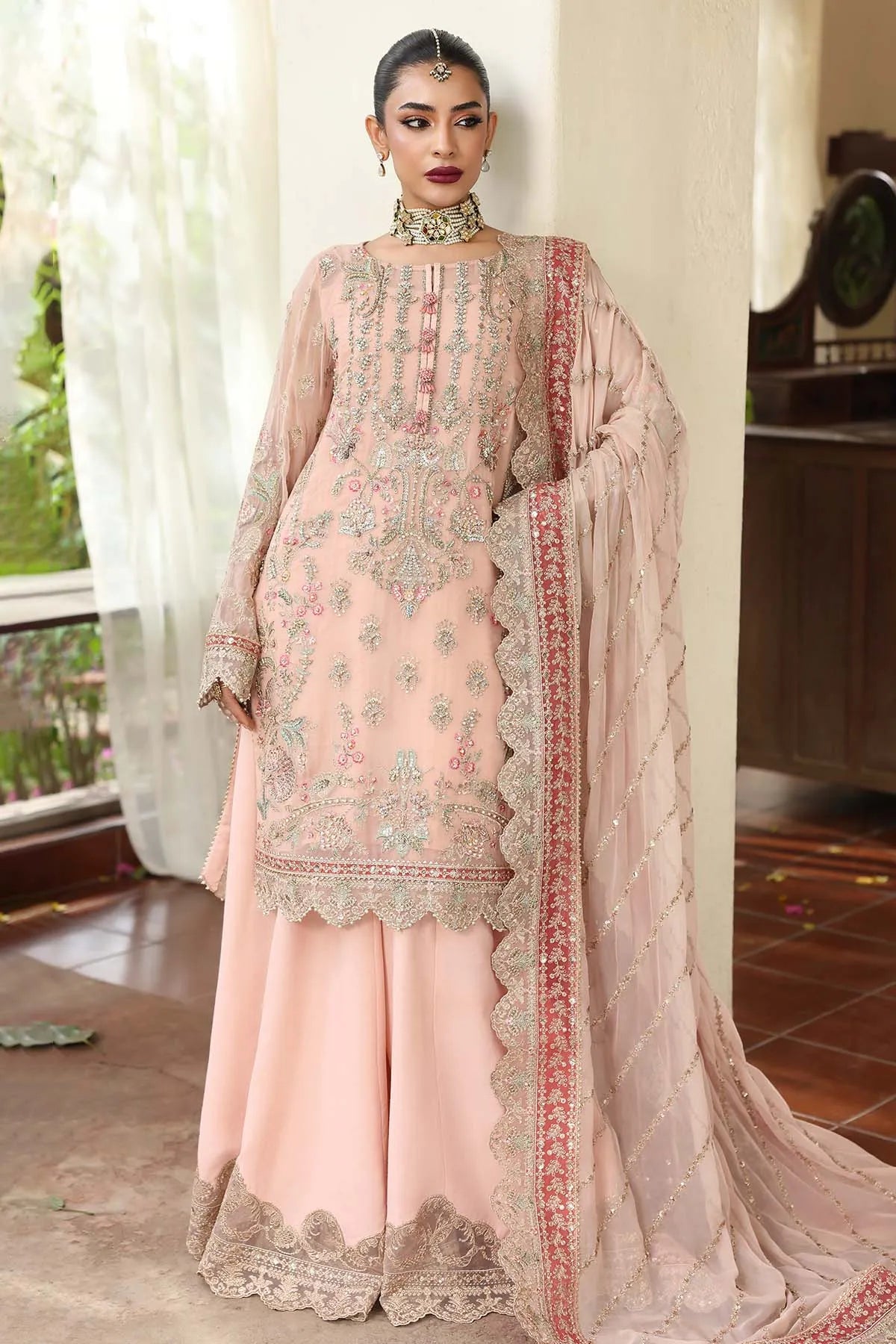 Woman wearing a traditional pink embroidered outfit with a matching dupatta indoors.