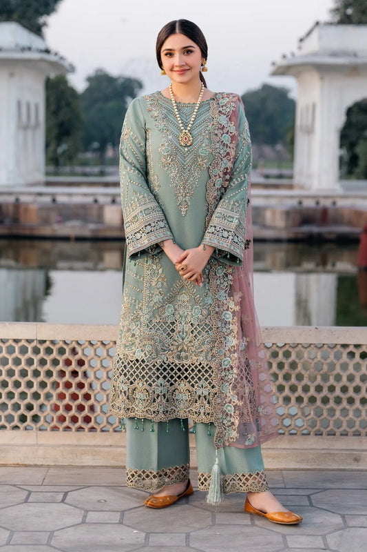 A woman wearing a green and pink traditional outfit with embroidery and an organza dupatta, standing near a water body.