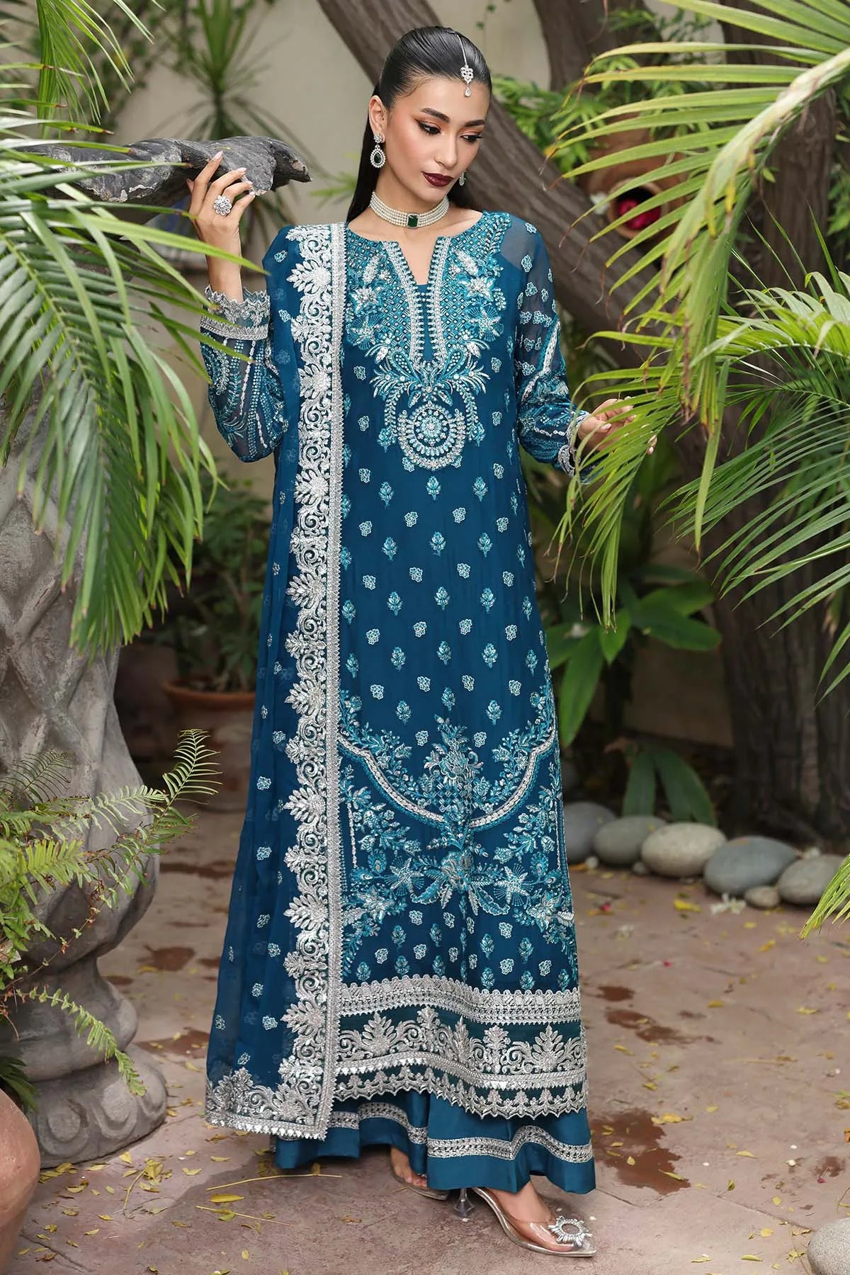 Woman in a blue embroidered traditional outfit standing outdoors with greenery.