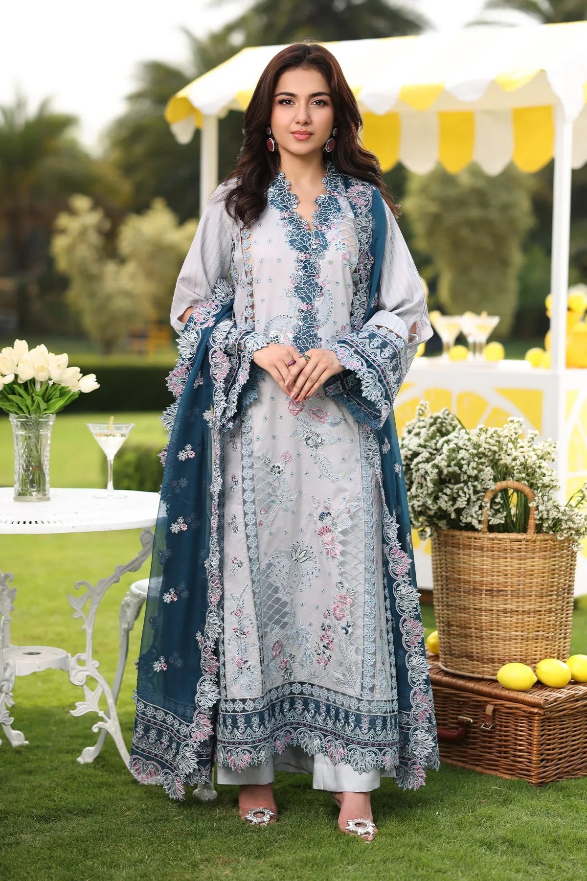 A woman wearing a traditional-style dress with floral embroidery, consisting of a shirt, dupatta, and trousers, in shades of teal and liver.